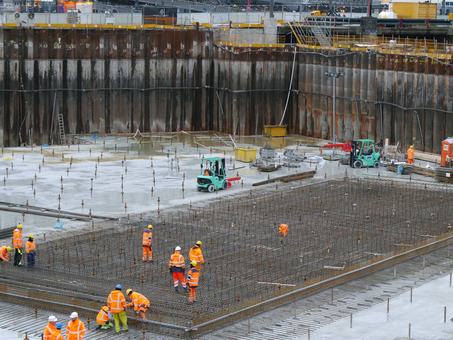 Stor byggegrop med spuntvegg og armeringsnett. Arbeidere i synlighetstøy spredt utover flaten.